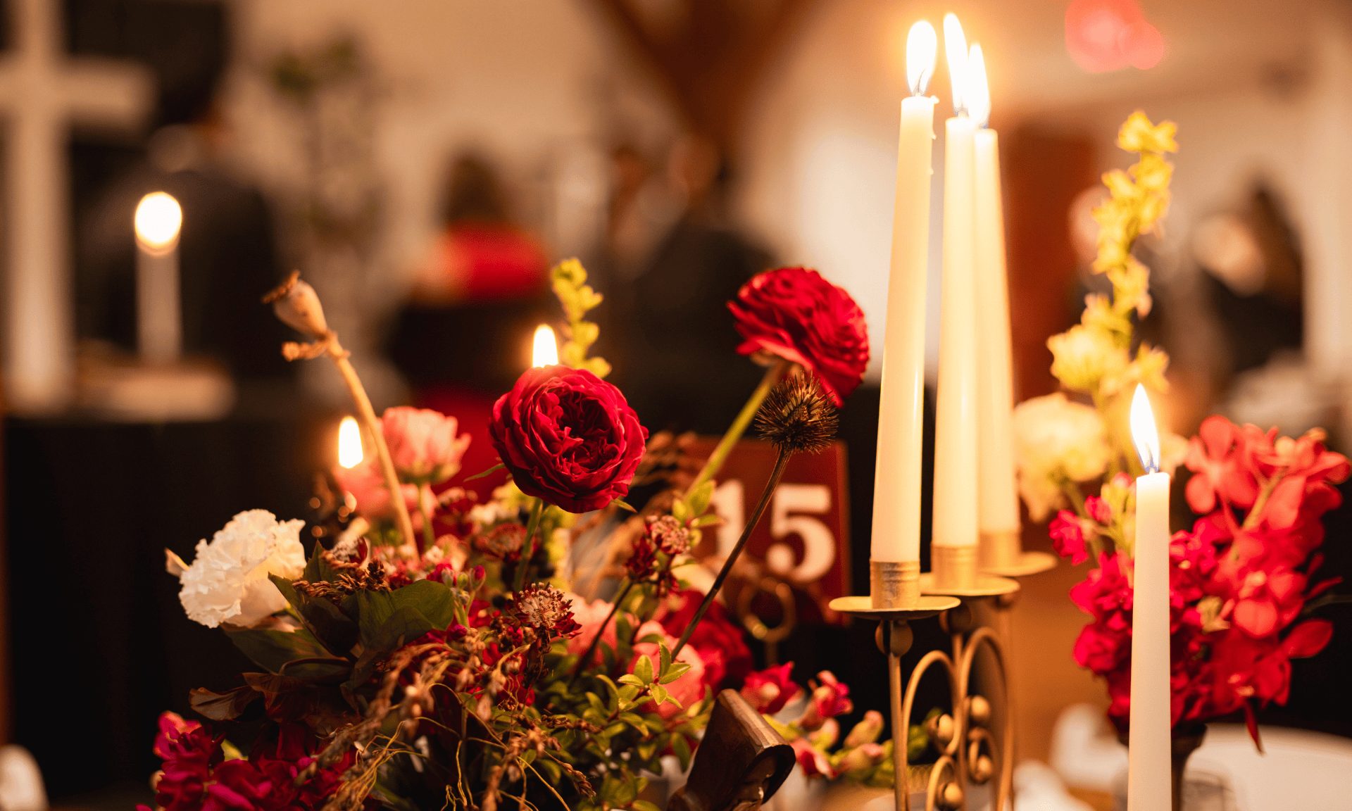 Deep red roses and lit taper candles beside a table number at a reception.