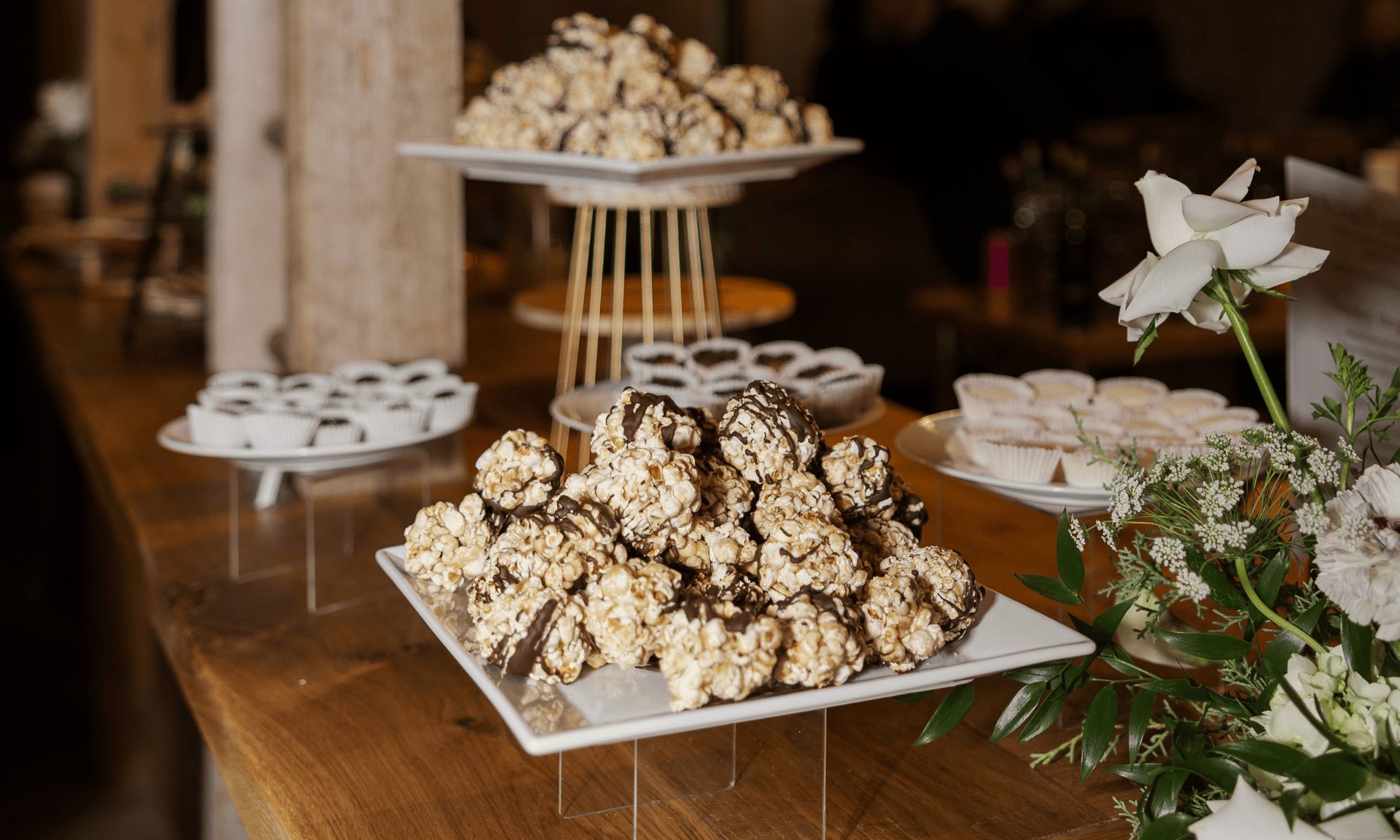 A dessert table of chocolate drizzled popcorn balls and cupcakes on white stands beside white roses.