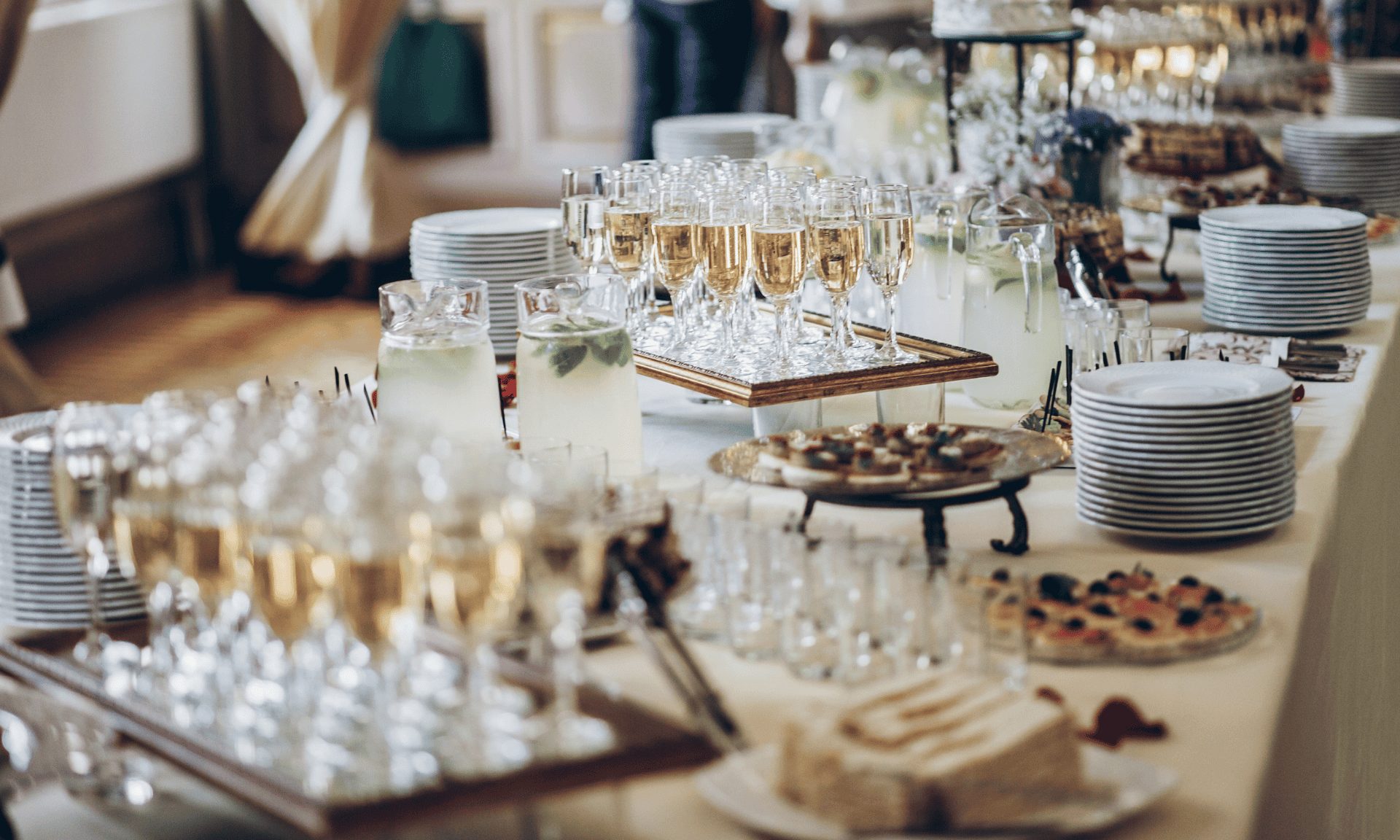 A long buffet table set with filled champagne flutes, stacked plates, glass pitchers and trays of canapes.