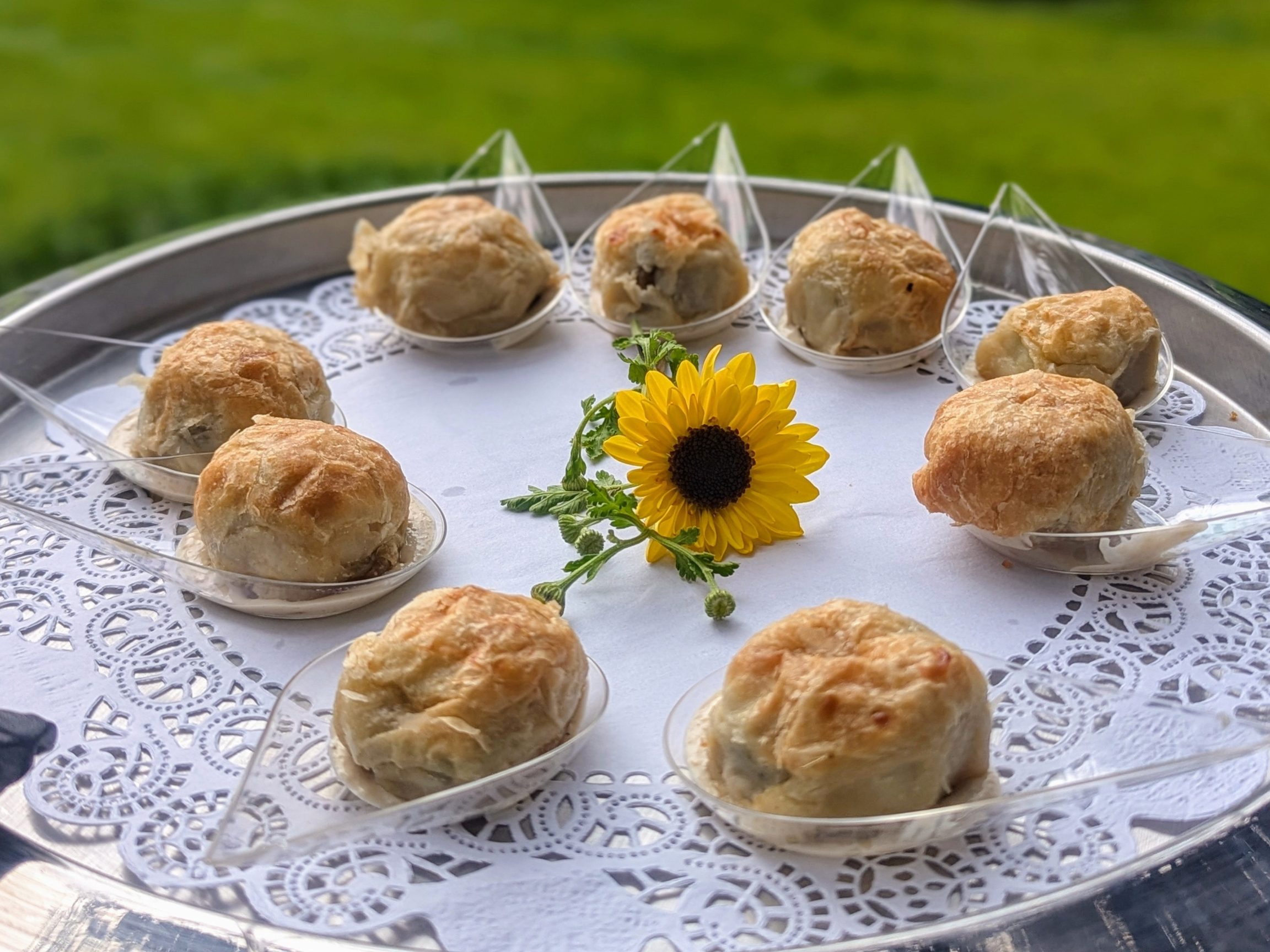 Puff pastry hors d'oeuvres on tasting spoons arranged around a sunflower on a silver tray.