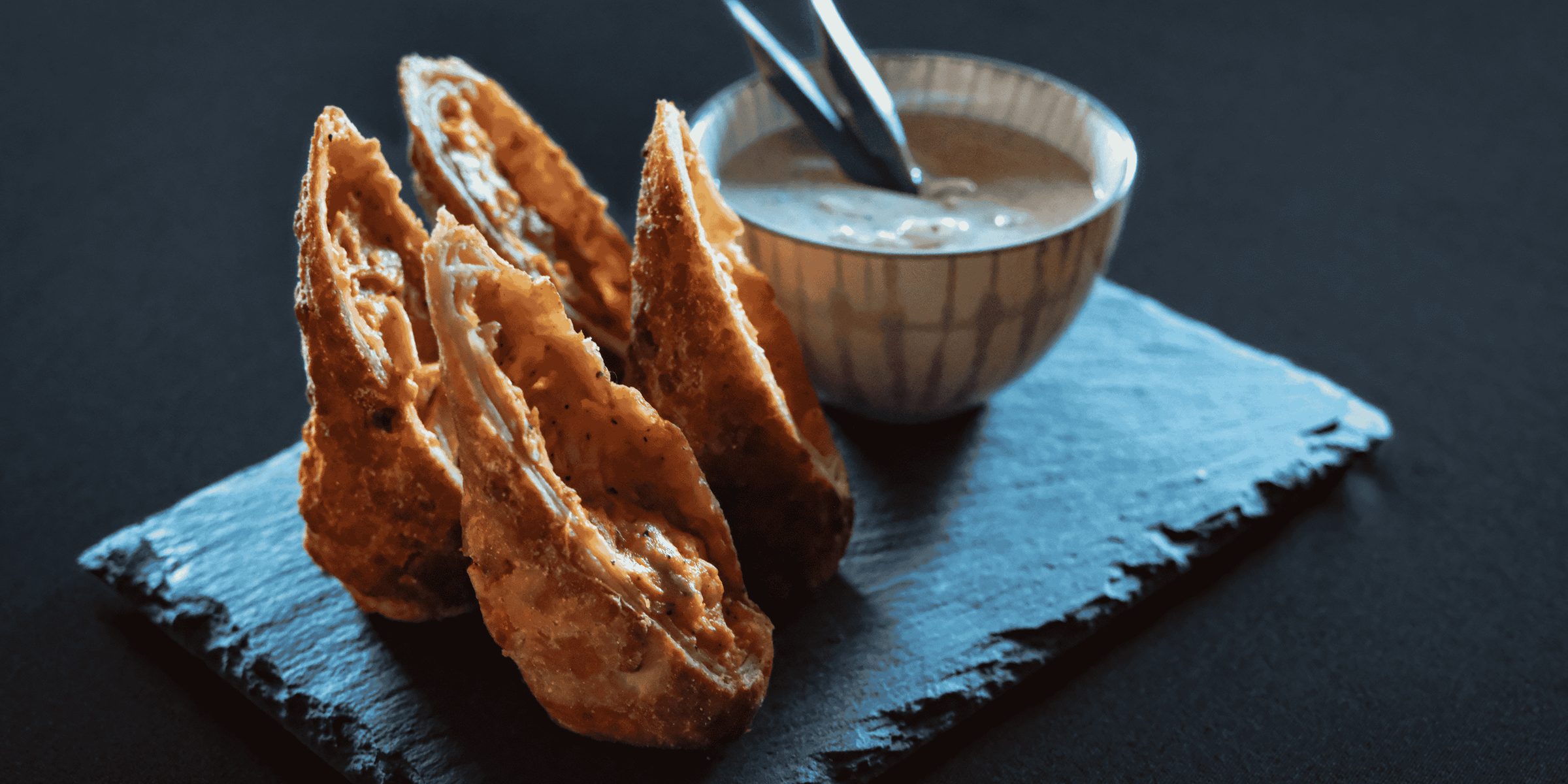 Crisp filled pastries fanned across a dark slate board beside a small bowl of dipping sauce.