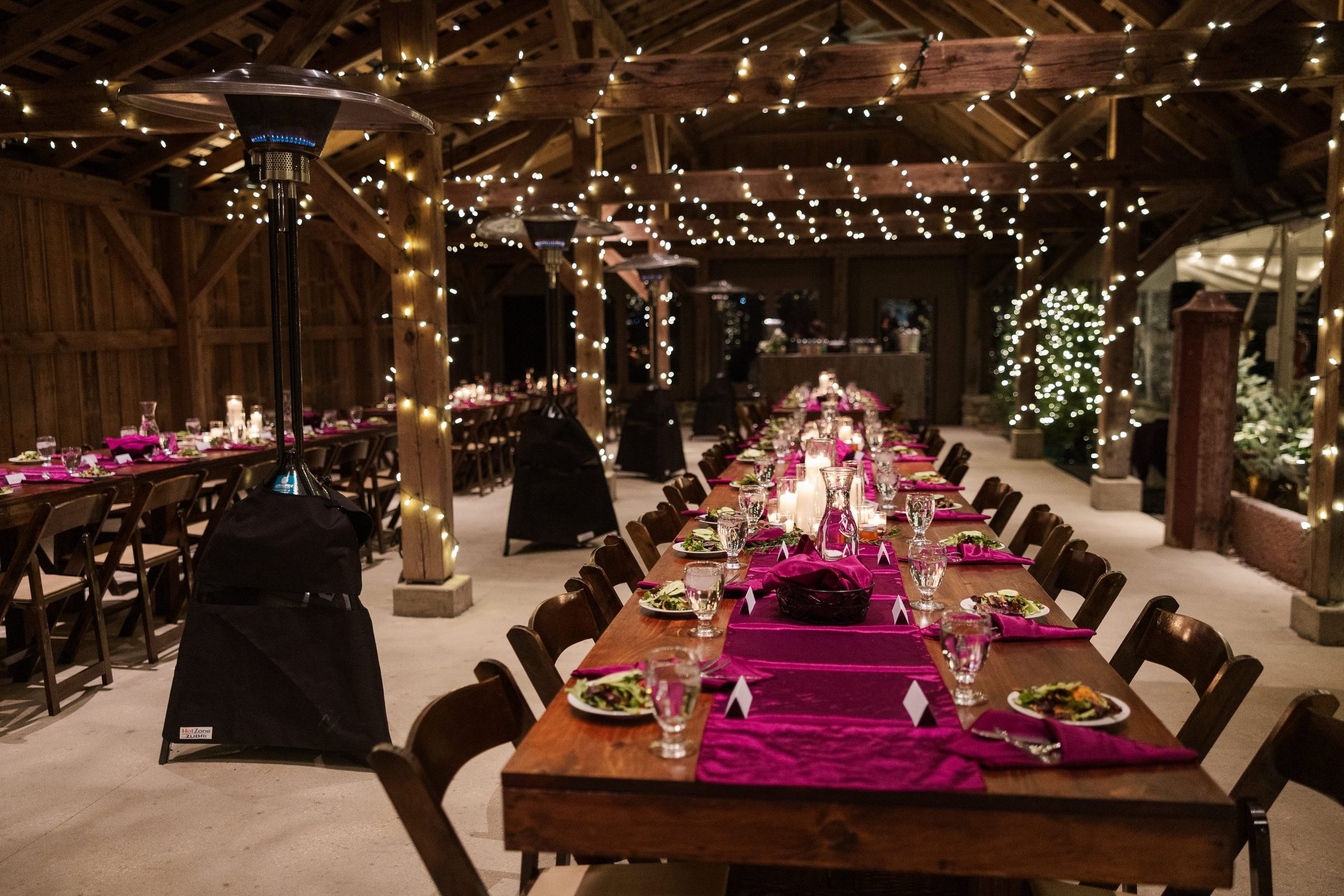 Long banquet tables set with magenta runners and candles in a string-lit barn.