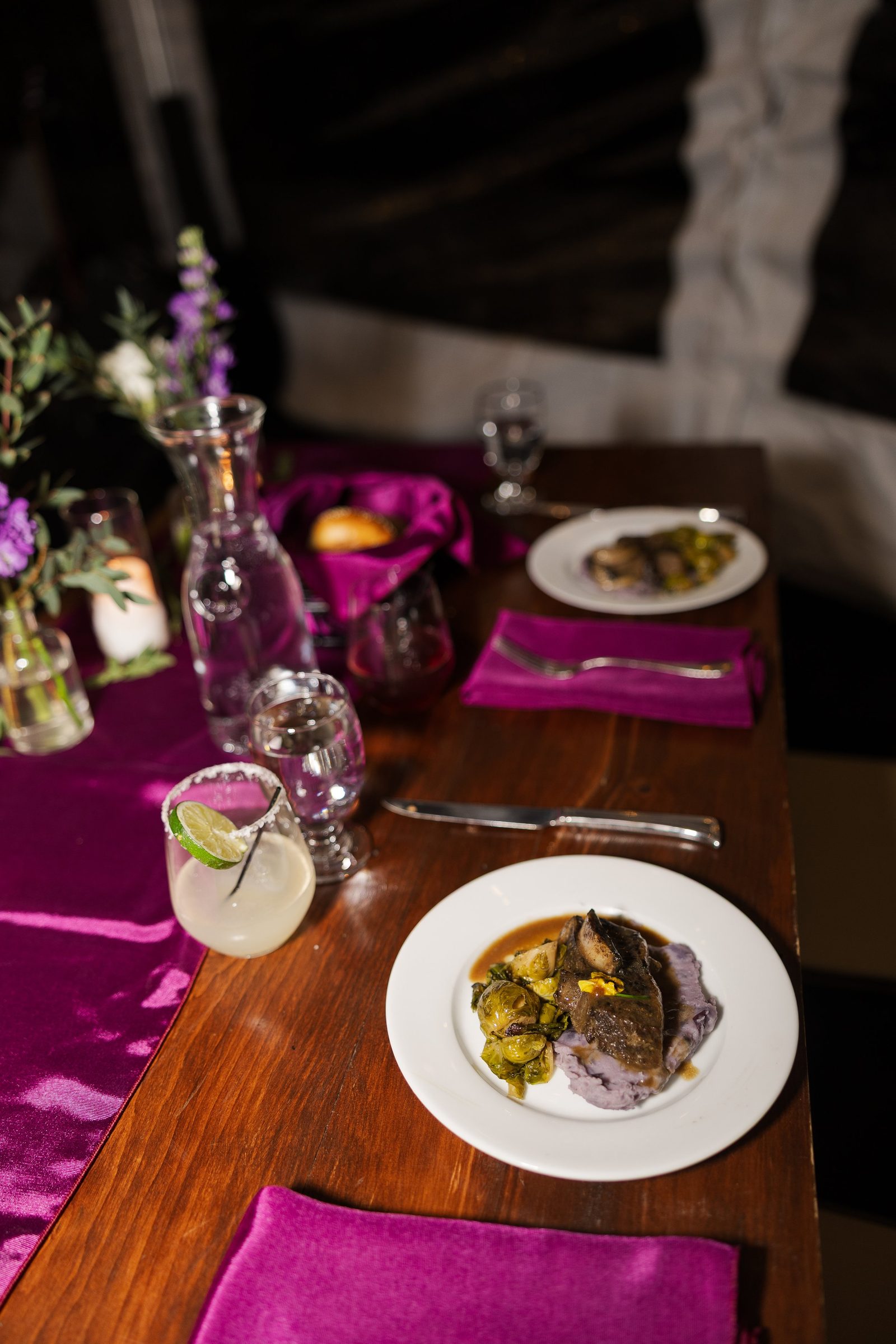 Two plated main courses on a wooden table dressed with magenta linens and glassware.
