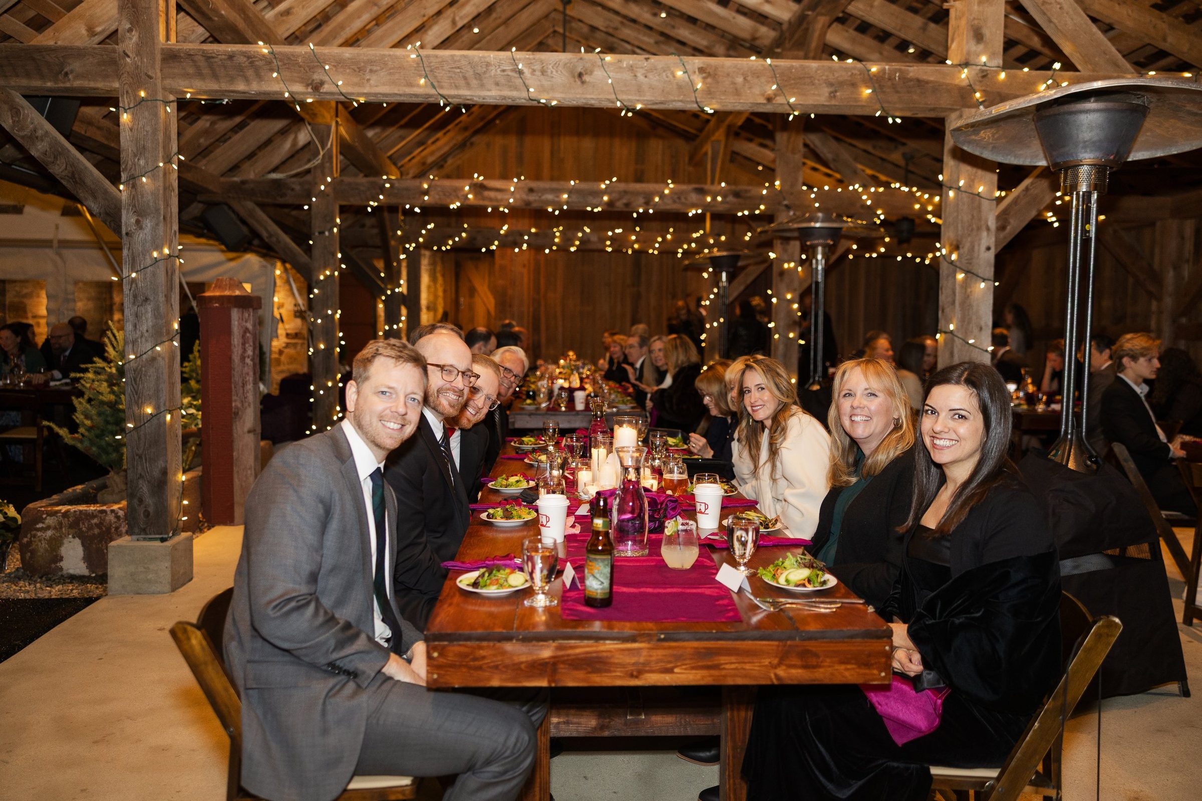 Guests seated along a long farm table under string lights in a timber-framed barn.