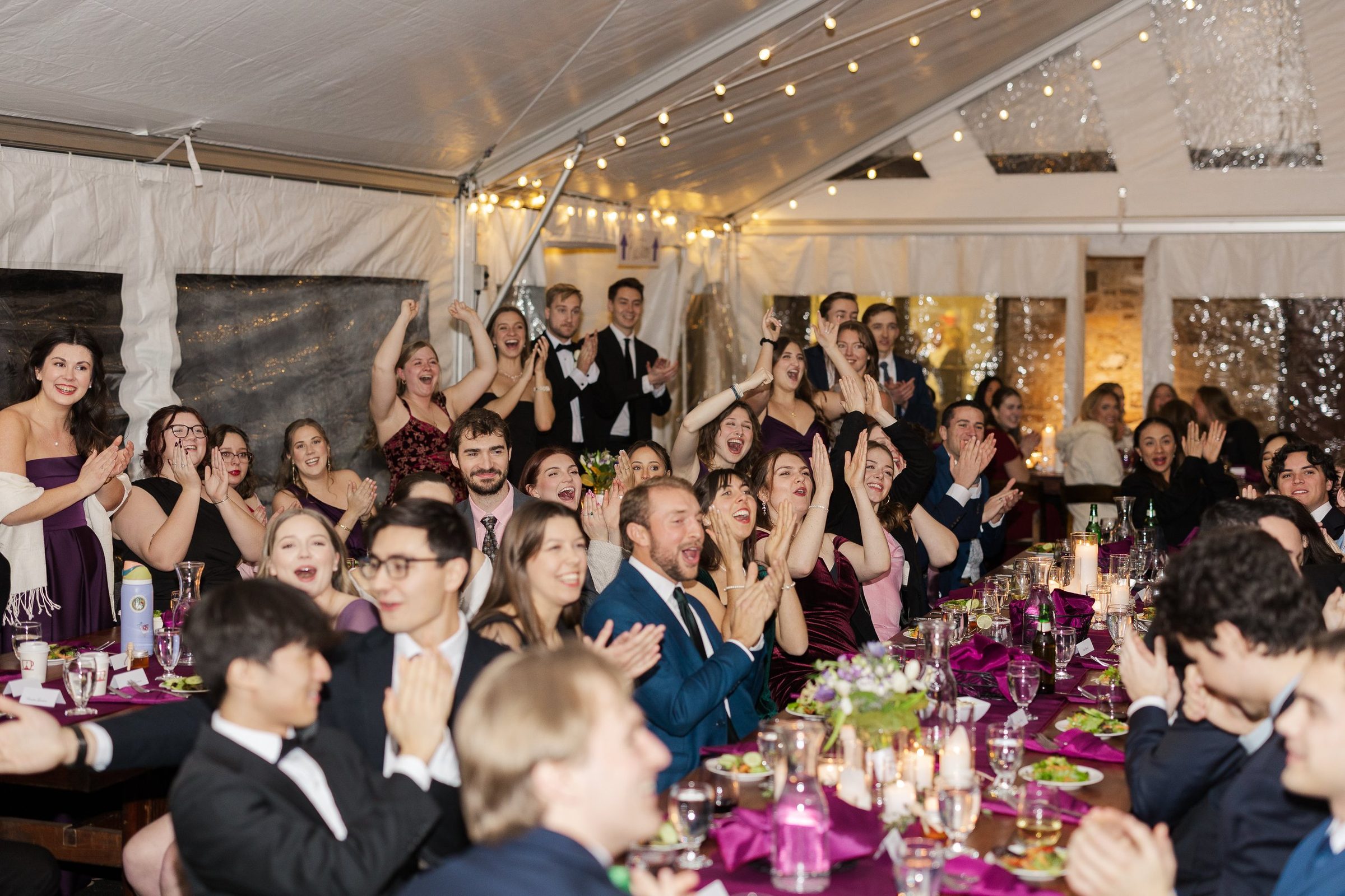 Wedding guests applauding at long tables under a tented reception with string lights.