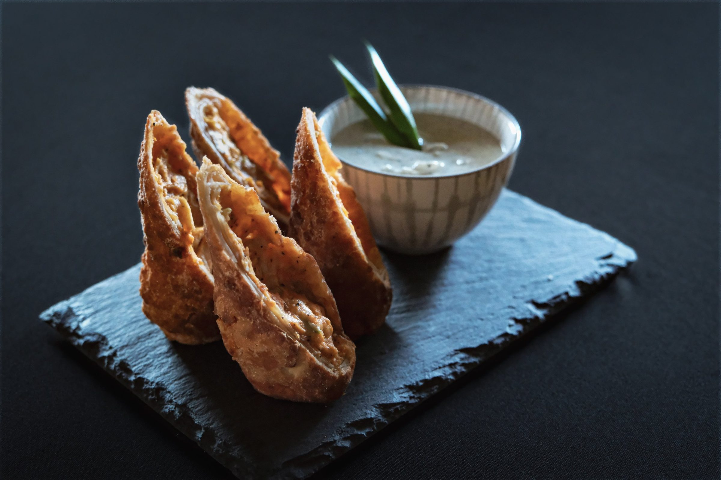 Four cheesesteak eggrolls stood upright on a slate board beside a bowl of dipping sauce.
