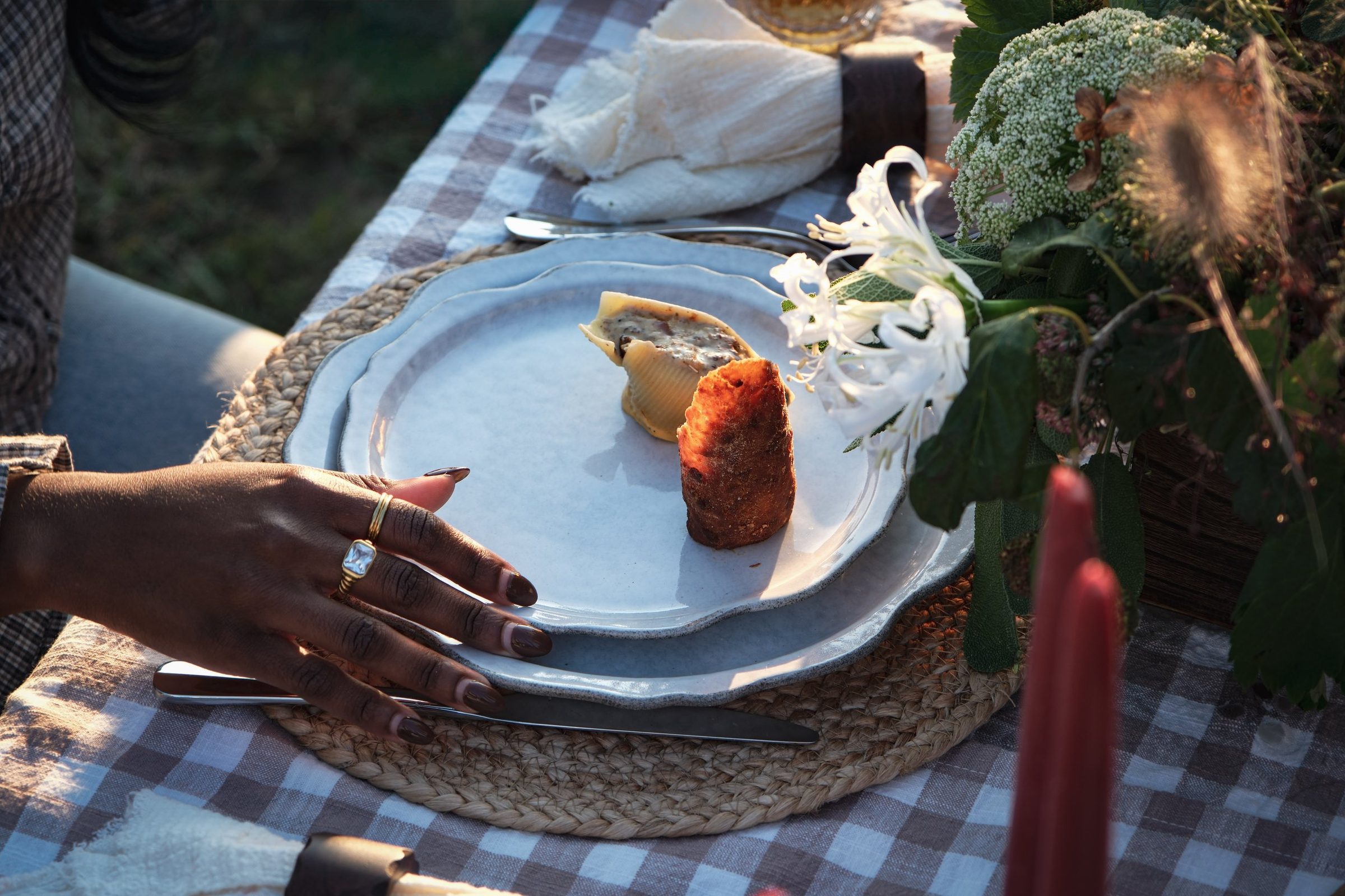 A hand reaching for a plate of stuffed pasta shell and a crisp croquette at an outdoor table set with checked linen.