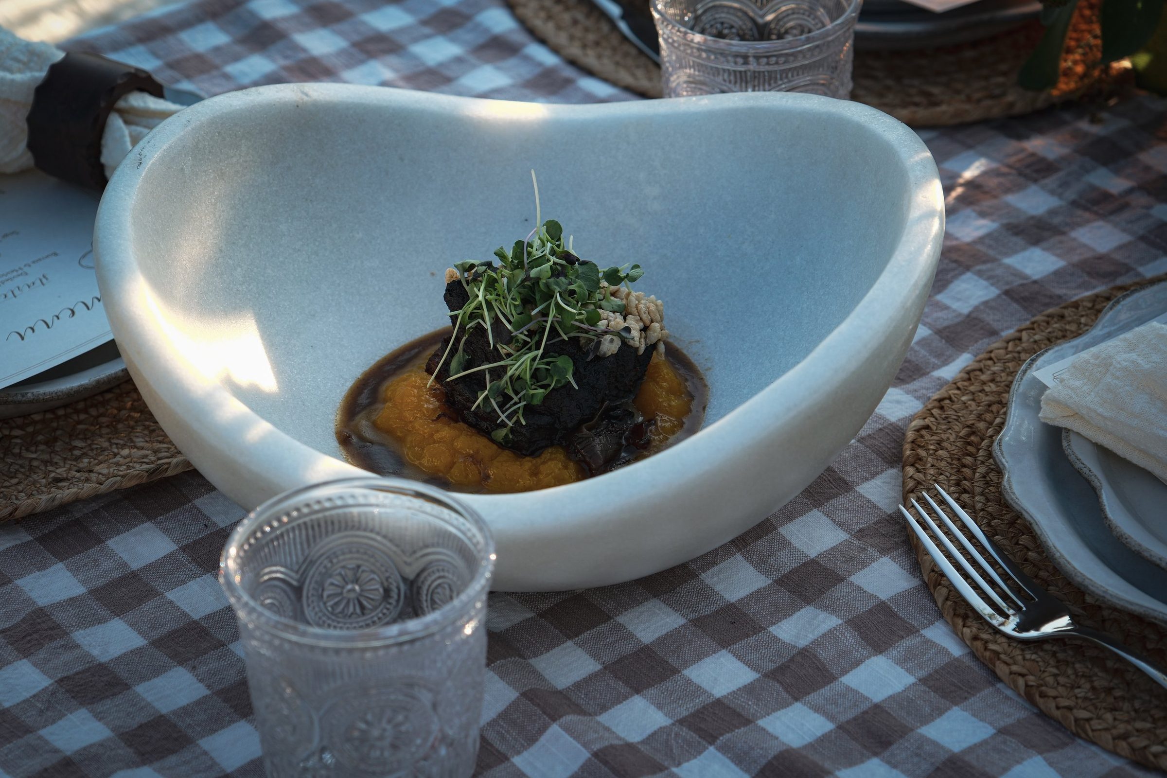 Braised short rib on squash puree, topped with microgreens, in a white bowl on a checked cloth.
