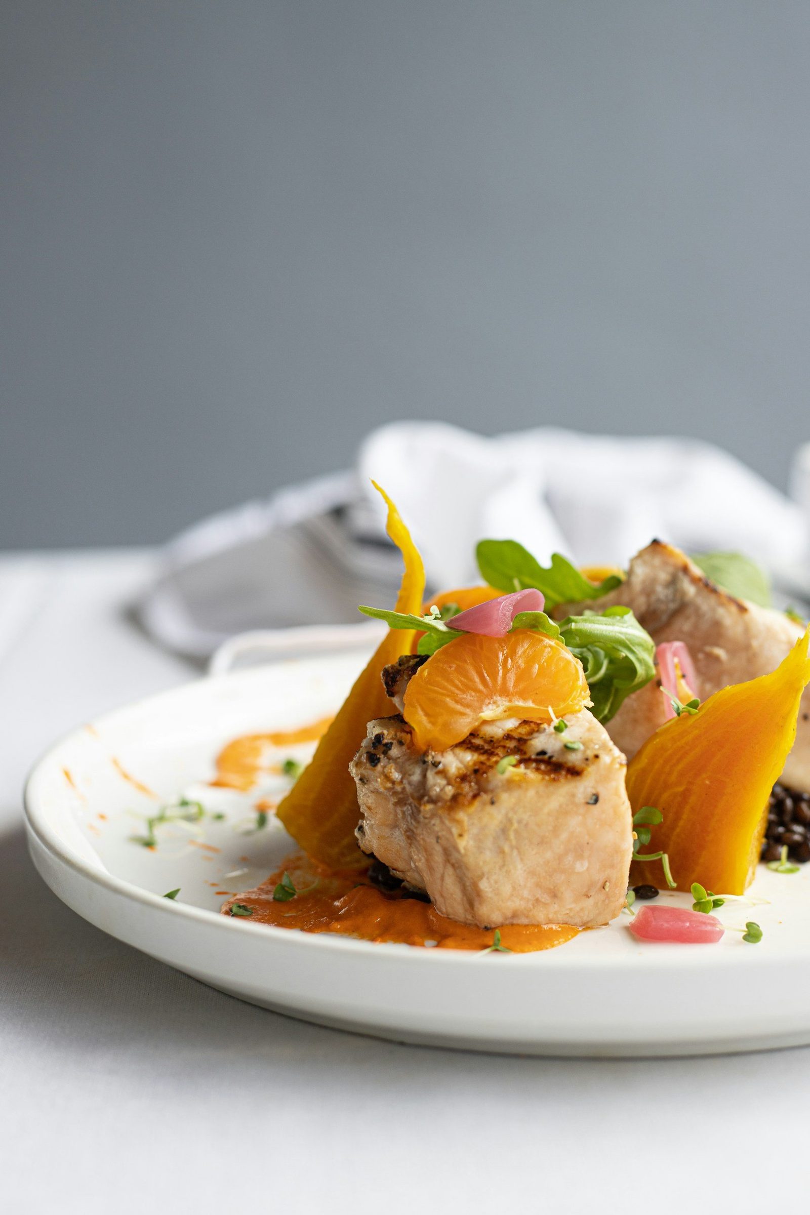 A seared fish fillet plated with golden beet, mandarin, pickled onion and a red pepper sauce.