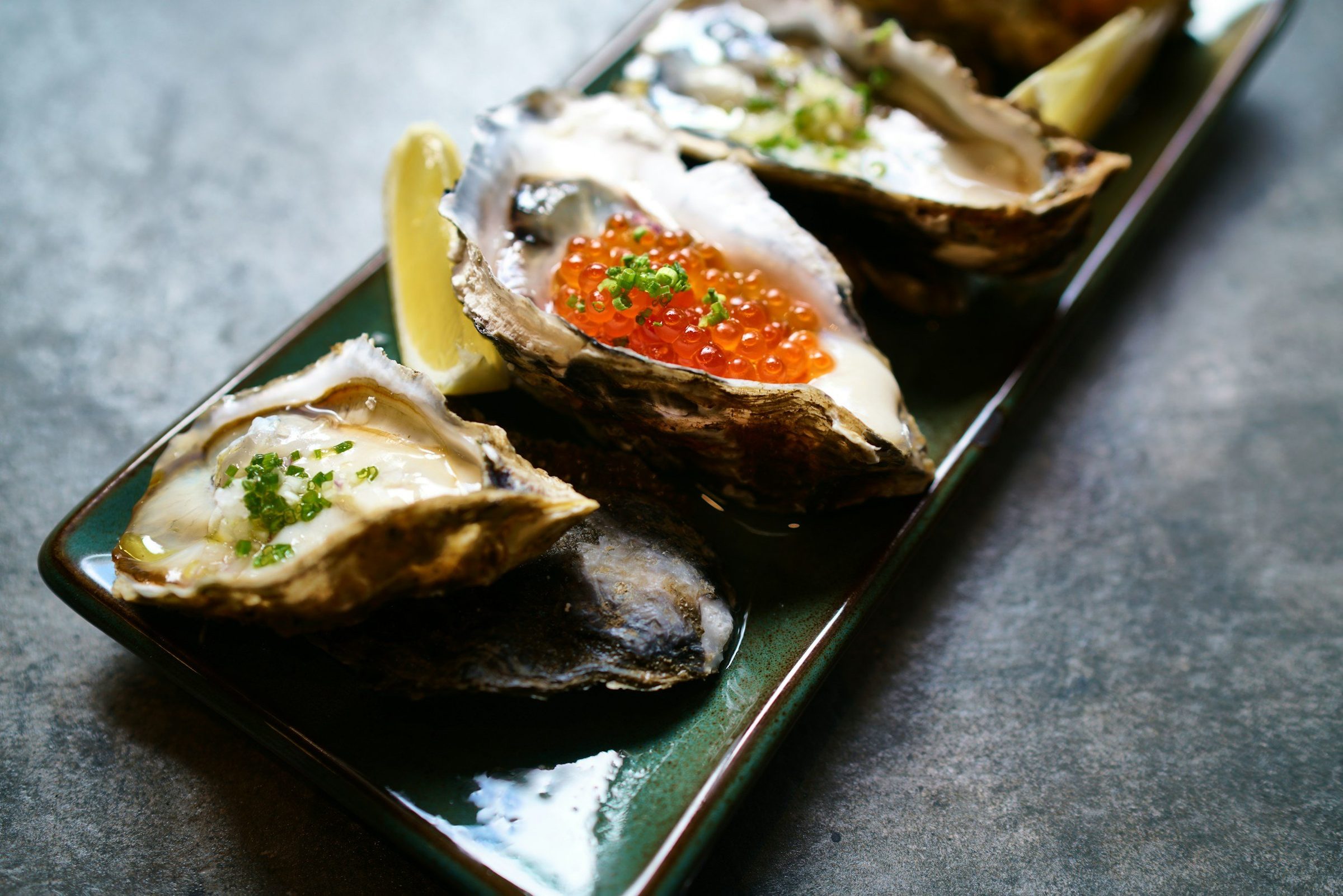 Oysters on the half shell on a dark green plate, one topped with salmon roe and chives, with a lemon wedge.