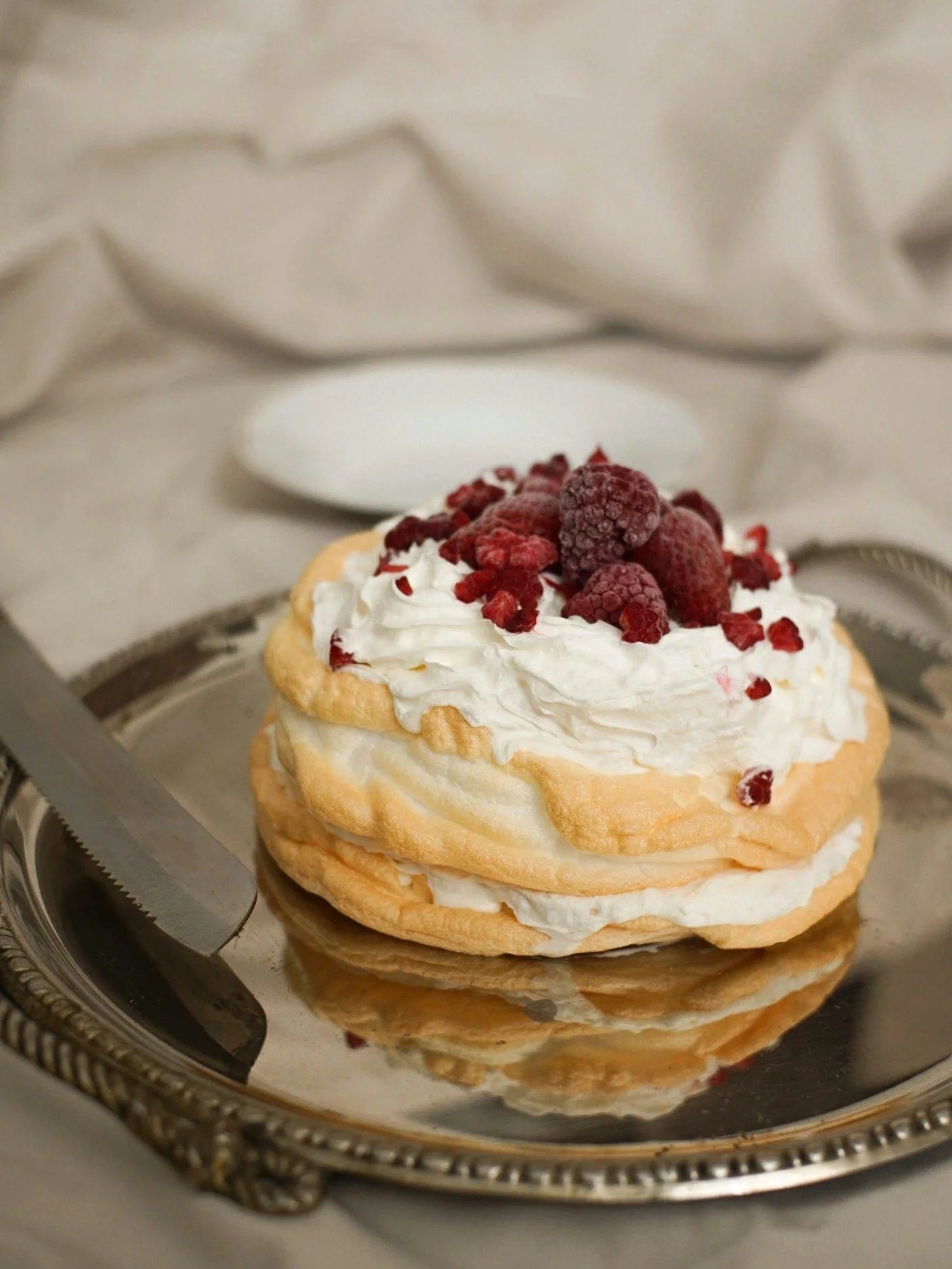 A layered meringue and cream dessert topped with raspberries on a silver tray.