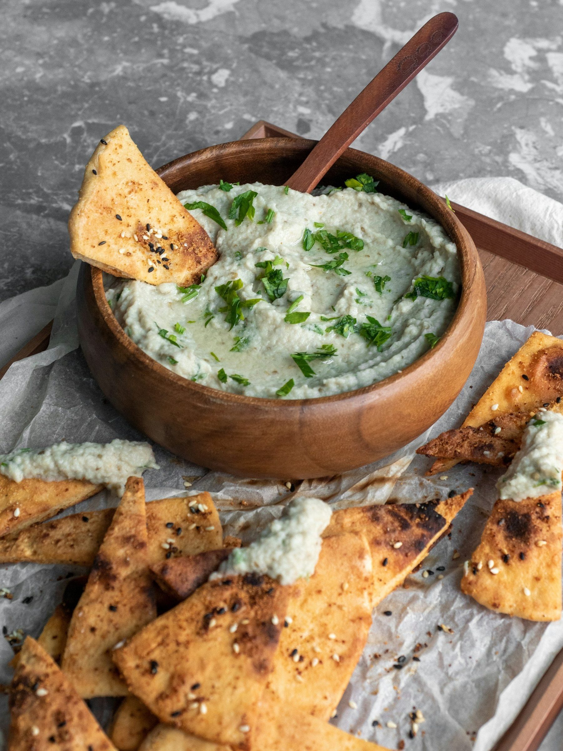 A wooden bowl of herbed white bean dip on a board with seeded pita chips.