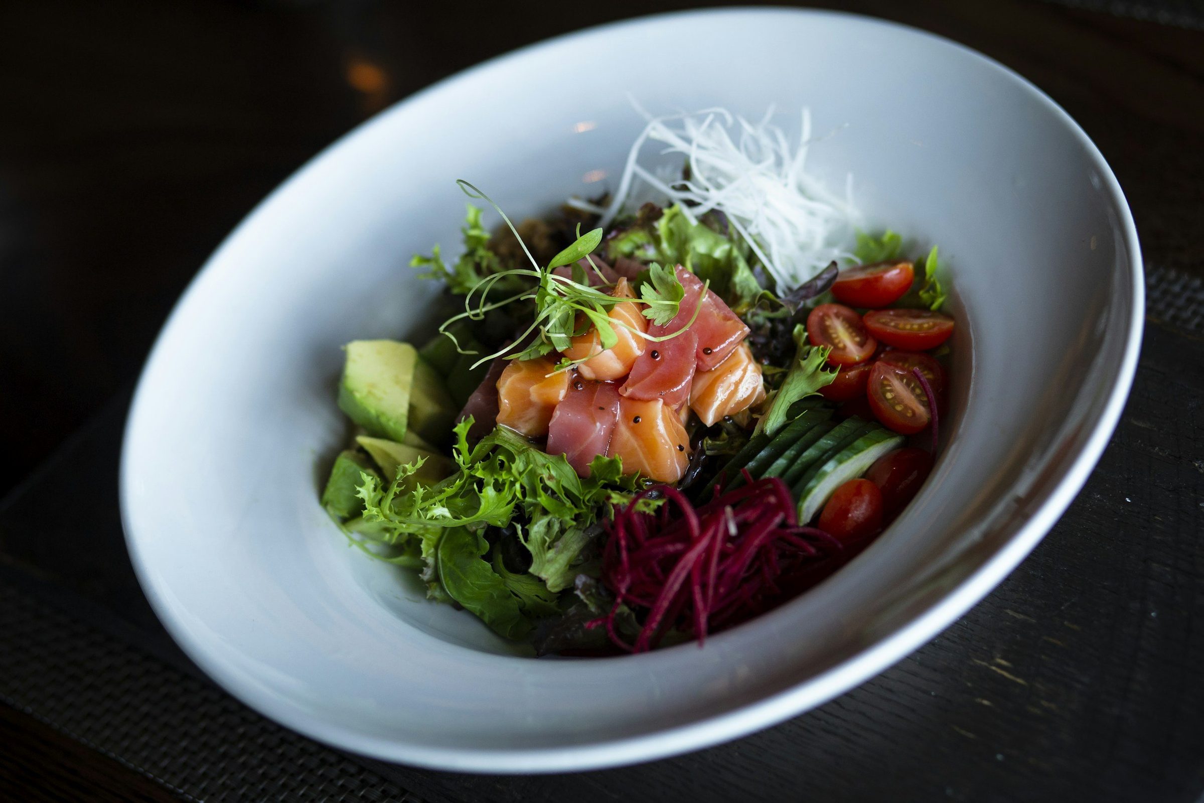 A white bowl of leaf salad topped with sliced cured salmon, avocado, cherry tomatoes and shredded beetroot.